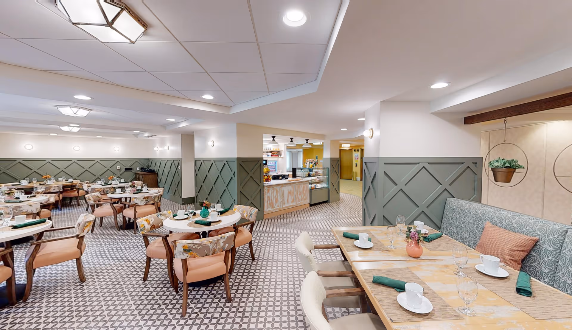 A bright and spacious dining room in a senior living facility with multiple round and rectangular tables set with white cups, plates, glassware, and green napkins. The room features patterned tile flooring, green wainscoting on the walls, cushioned chairs with floral upholstery, and a small counter area in the background with a display case. Ceiling lights provide ample illumination.