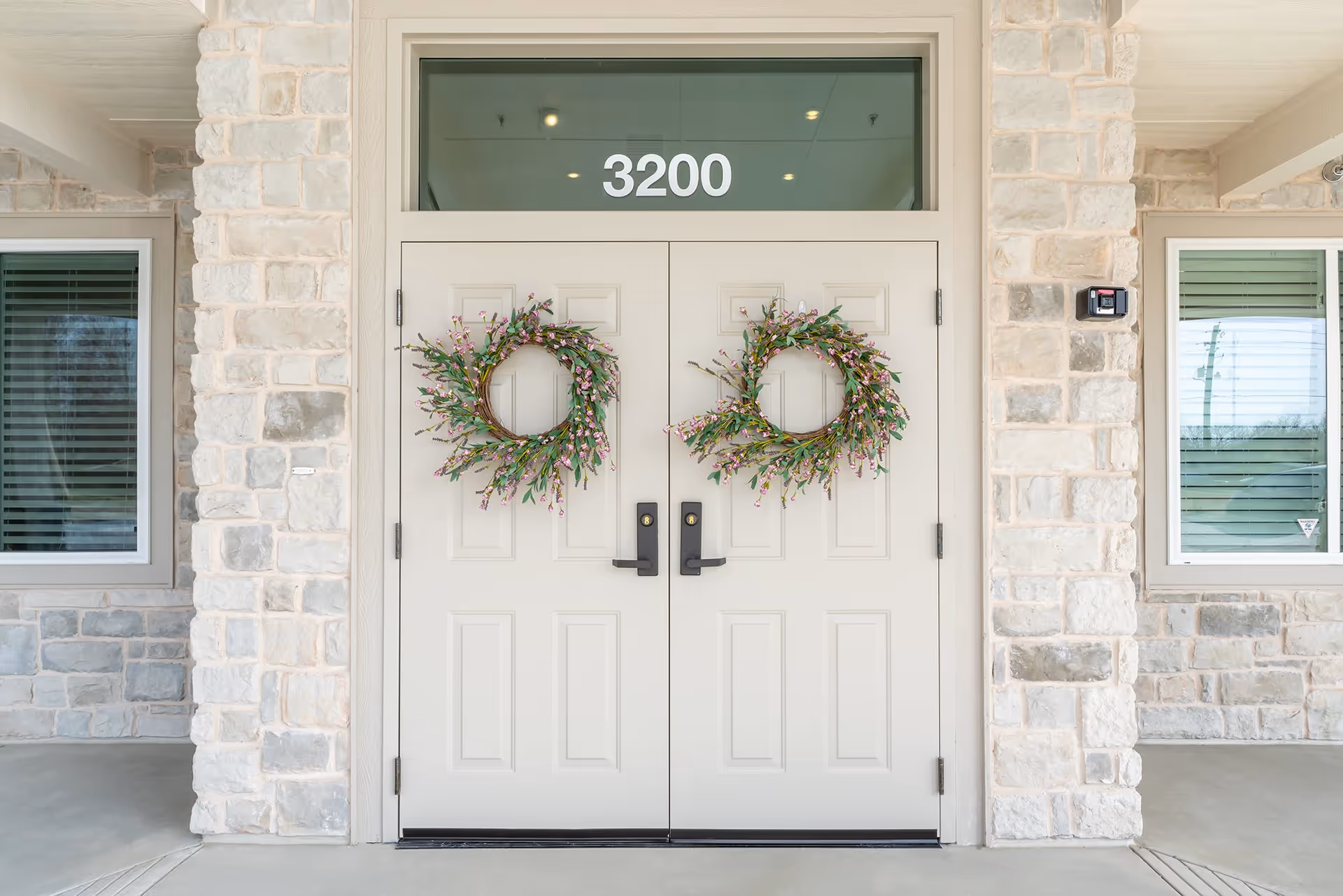 Double beige entrance doors with black handles, each decorated with a wreath of green leaves and small pink flowers. Above the doors is a transom window displaying the number 3200. The doors are set in a stone exterior wall with two windows on either side.