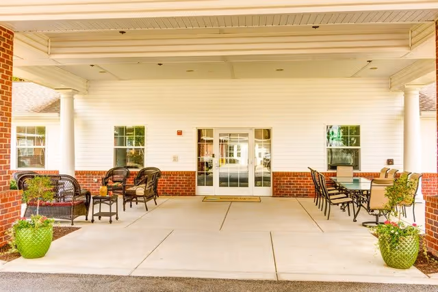 Covered outdoor seating area at the entrance of a building with white siding and brick accents. The area features two sets of seating arrangements: on the left, black wicker chairs with cushions and a small table, and on the right, a glass-top table with several chairs. Two large green planters with plants are placed on either side of the seating area.