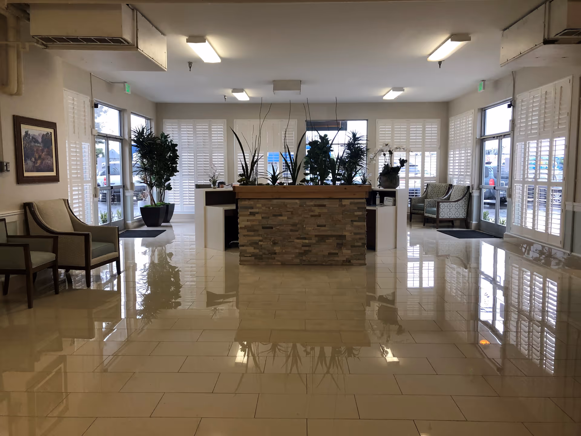 Bright polished lobby with a central stone-faced reception desk, potted plants, seating and large shuttered windows.