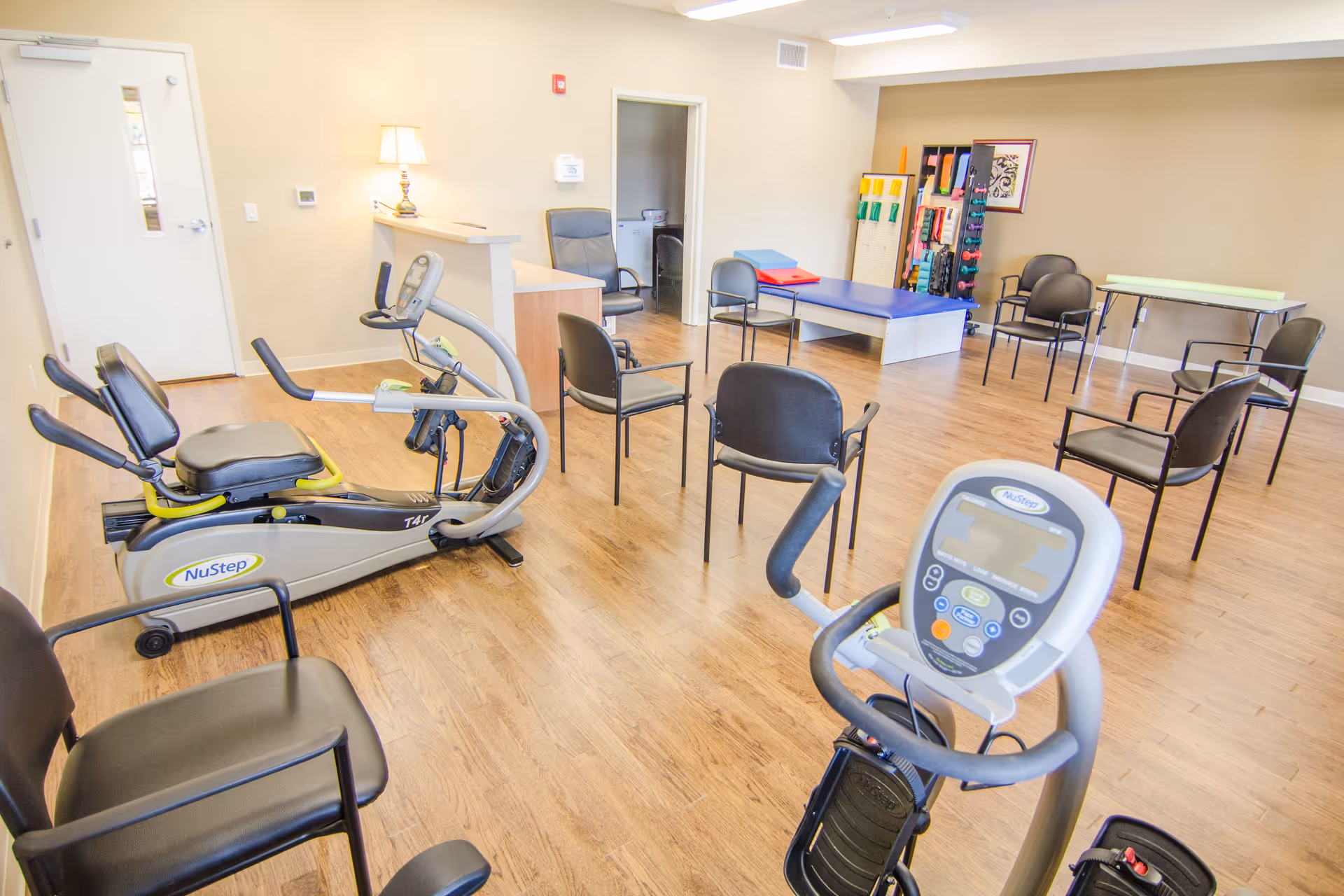 Bright exercise/therapy room with NuStep machines, chairs, mats, and rehabilitation equipment on a wood floor.