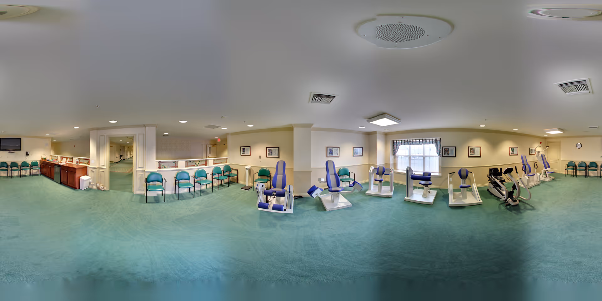 A spacious exercise room with green carpet and cream-colored walls. The room contains several pieces of exercise equipment with blue padding arranged in a row near a window with a valance. There are multiple green chairs lined up along the walls, a flat-screen TV mounted on one wall, and framed pictures hanging on the walls. The ceiling has recessed lighting and ventilation fixtures.