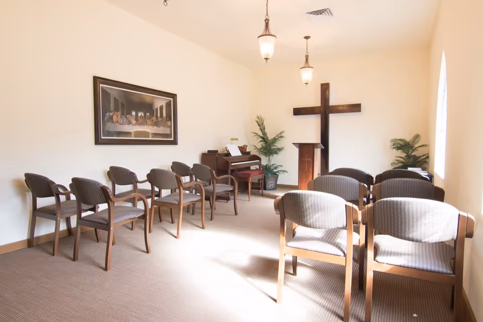A small chapel room with rows of wooden chairs arranged facing a wooden podium and a large wooden cross mounted on the wall. There is a piano in the corner, two potted plants, and a framed picture of The Last Supper on the wall. The room is softly lit by two hanging light fixtures and natural light from a window.