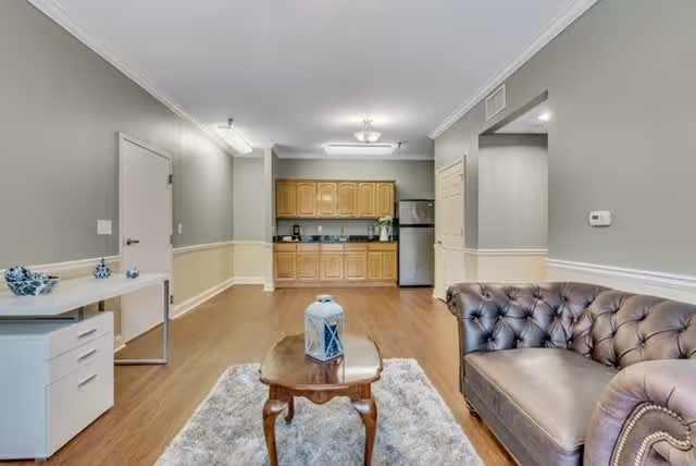 A spacious interior room featuring a small kitchen area with wooden cabinets, a stainless steel refrigerator, and a coffee maker. In the foreground, there is a brown leather tufted sofa and a wooden coffee table with a decorative lantern on a light-colored rug. To the left, a white desk with drawers and decorative items is placed against the wall. The room has light gray walls with white trim and wood flooring.