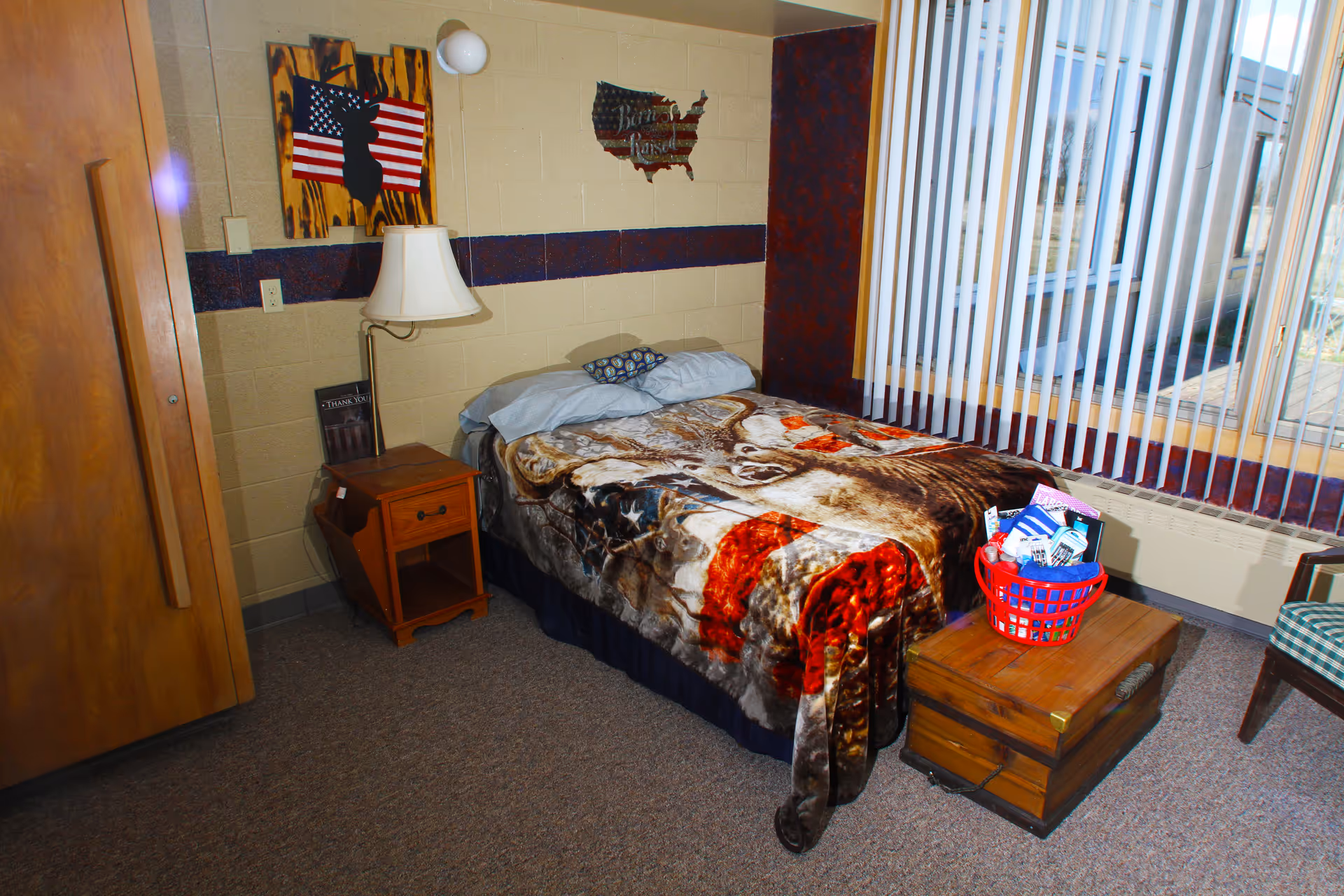A small bedroom with a single bed covered by a blanket featuring a deer design. Next to the bed is a wooden nightstand with a lamp and a book. On the wall above the nightstand are two decorations, one with an American flag and a silhouette of a rabbit, and another shaped like the United States with text. A large window with vertical blinds lets in natural light. A wooden chest with a red basket filled with various items sits at the foot of the bed. The room has carpeted flooring and a wooden door on the left side.