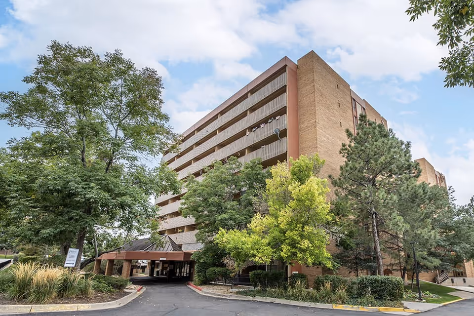 Exterior view of a multi-story brick senior living facility building surrounded by trees and landscaping under a partly cloudy sky. The entrance has a covered driveway with a sign near the sidewalk.
