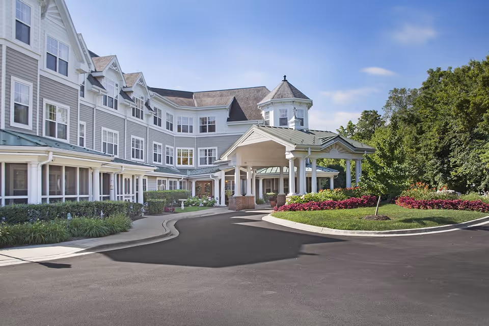 Front exterior of a multi-story senior living building with a covered porte-cochere, landscaped roundabout, and clear sky.