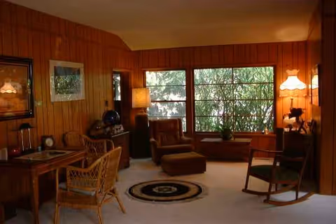 Cozy wood-paneled living room with chairs, an ottoman, and large windows.