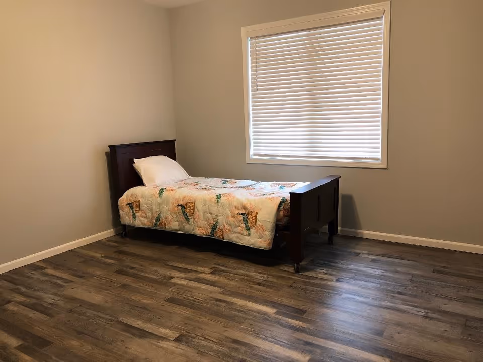 Simple bedroom with a single wooden bed near a window with closed blinds and wood-look flooring.