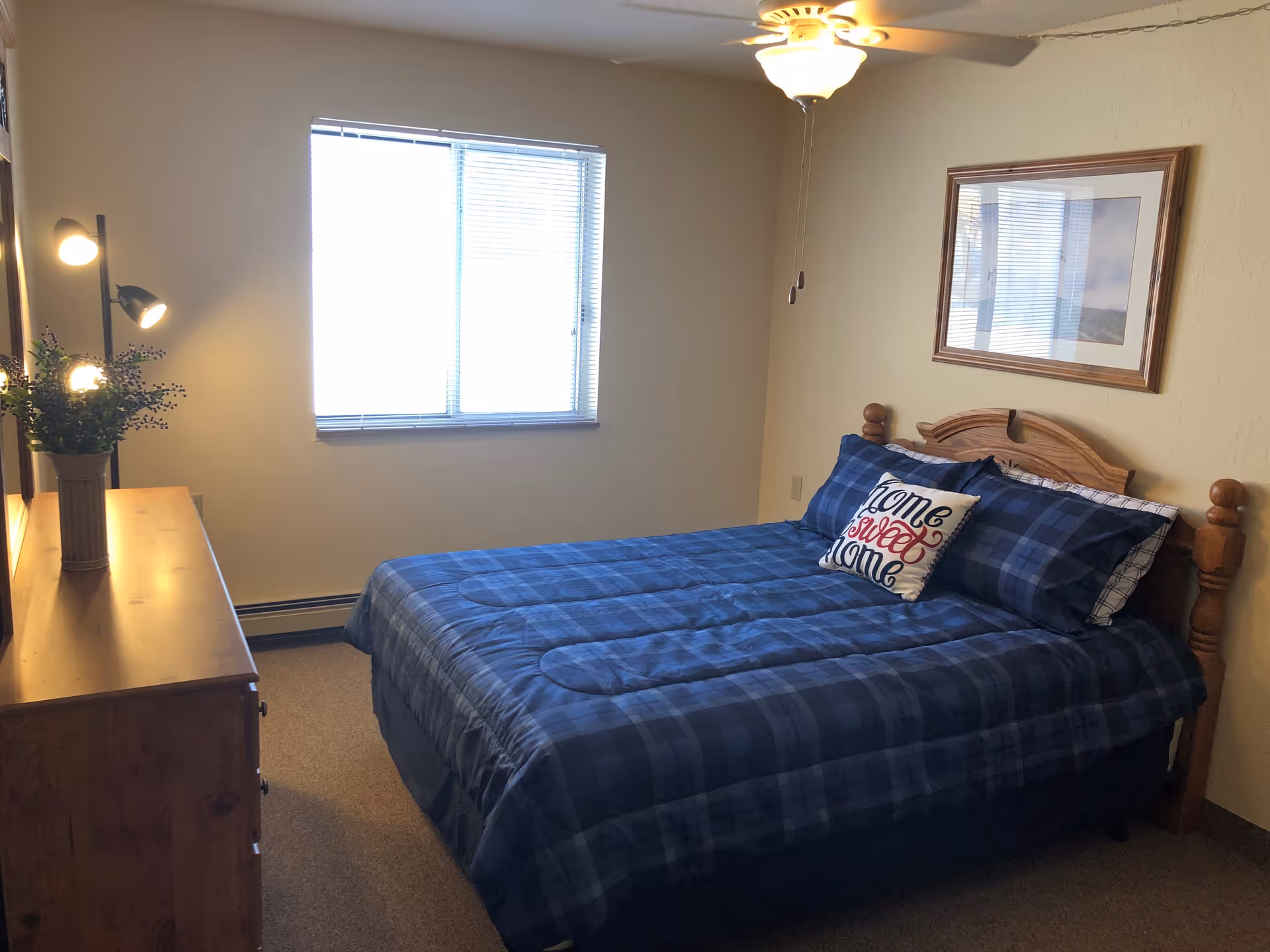 A bedroom with a wooden bed frame and a blue plaid comforter. There are two pillows, one with the text 'home sweet home'. A wooden dresser with a vase of flowers and a standing lamp with three lights is on the left side. A window with blinds is centered on the back wall, and a framed picture hangs above the bed. A ceiling fan with a light is on the ceiling.