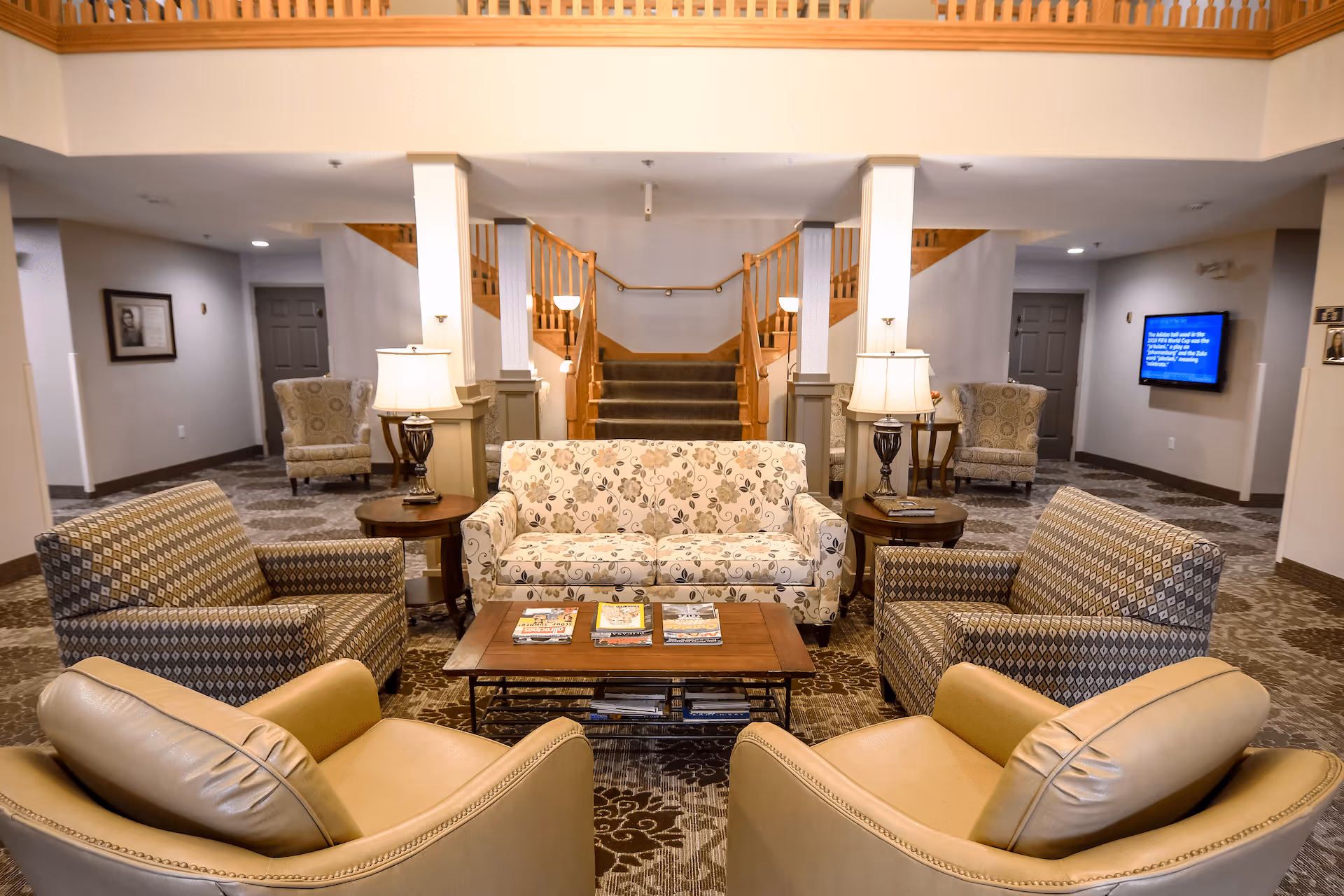 A cozy senior living facility common area with a floral patterned sofa, two patterned armchairs, and two beige leather chairs arranged around a wooden coffee table with magazines. Two table lamps sit on side tables beside the sofa. In the background, a staircase with wooden railings leads to an upper floor, and additional armchairs and a wall-mounted TV are visible.