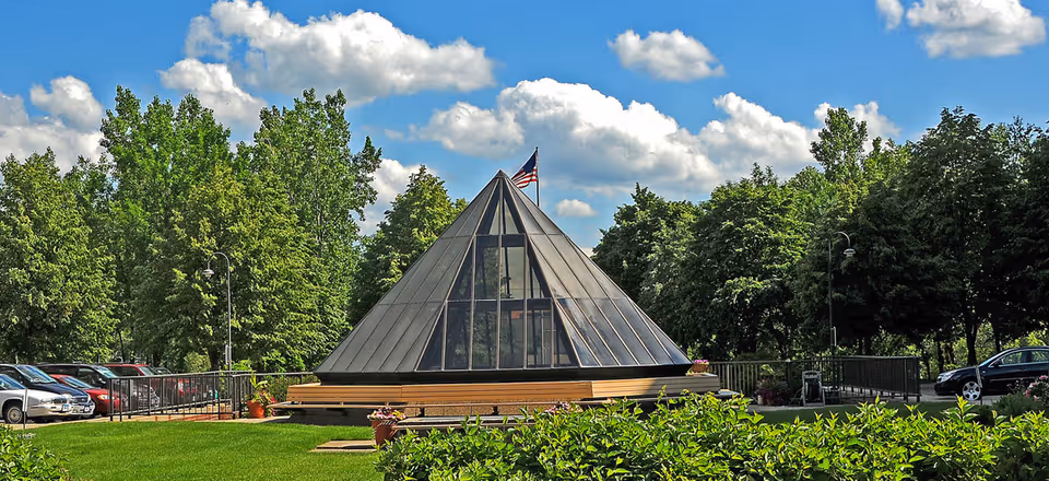 Pyramid-shaped glass pavilion with an American flag on top on a manicured lawn, surrounded by trees and parked cars.