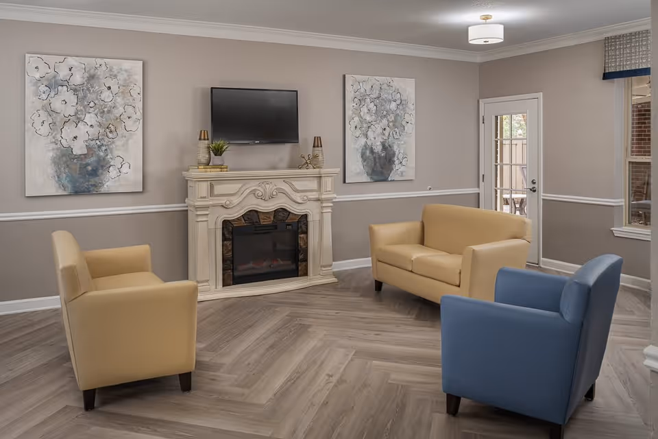 A cozy living room with a beige loveseat, two beige armchairs, and one blue armchair arranged around a decorative white fireplace with a mounted flat-screen TV above it. The walls are painted light gray with white trim, and two large floral paintings hang on either side of the fireplace. A door with glass panes and a window provide natural light to the room.