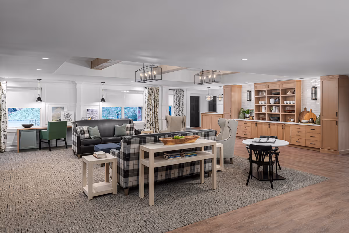 A spacious and well-lit living area in Richmond Place featuring a mix of comfortable seating including a plaid sofa, a black leather sofa, and two beige wingback chairs. There is a wooden console table with books and a decorative bowl of green apples. The room has large windows with patterned curtains, a small desk with a green chair, and built-in wooden cabinetry along one wall displaying dishes and decorative items. Modern light fixtures hang from the ceiling, and the floor is a combination of carpet and wood.