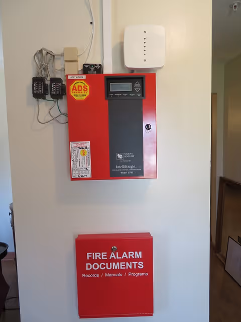 Red fire alarm control panel and a box labeled 'FIRE ALARM DOCUMENTS' mounted on a white interior wall.