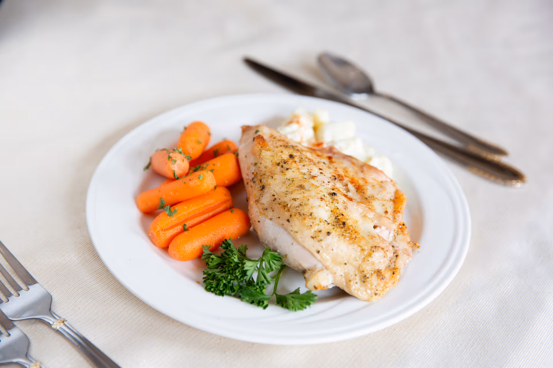 A white plate with a serving of cooked carrots, a piece of seasoned grilled chicken breast, and a garnish of parsley, placed on a light-colored tablecloth with a fork, knife, and spoon nearby.