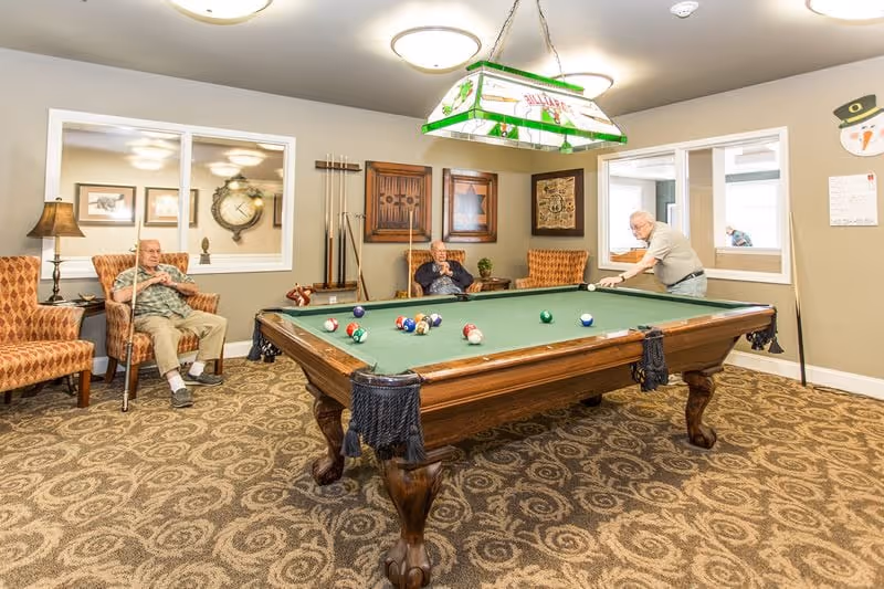 Three elderly men in a room with a pool table. One man is playing pool while the other two sit in armchairs holding pool cues. The room has patterned carpet, framed artwork on the walls, and a hanging light fixture above the pool table.
