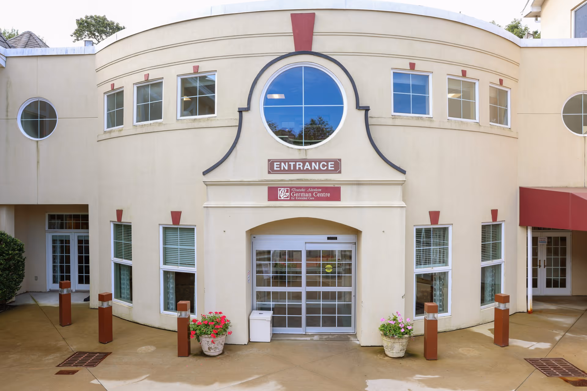 Front entrance of the German Centre Home building with a curved facade, multiple windows, a circular window above the entrance, and two flower pots with pink and red flowers on either side of the automatic sliding glass doors.