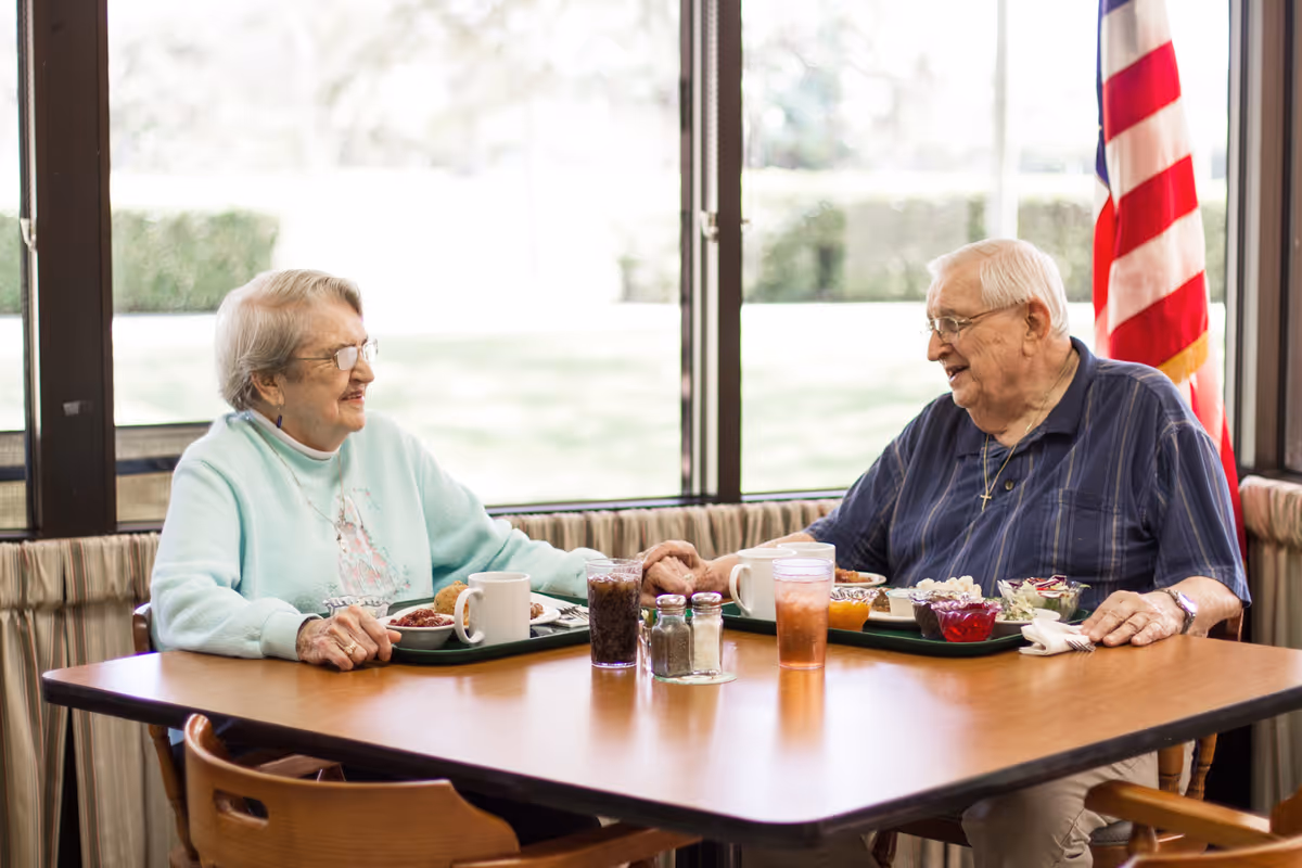 An elderly woman and man sitting at a dining table in a bright room with large windows. They are holding hands and smiling at each other. Each has a tray with food and drinks in front of them. An American flag is visible in the background.