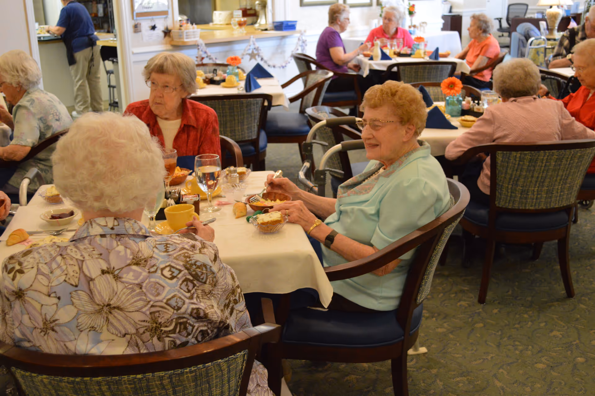 Several elderly women sitting at tables in a dining room, eating and conversing. The room has a cozy atmosphere with floral decorations and a carpeted floor. A staff member is visible in the background near a counter.