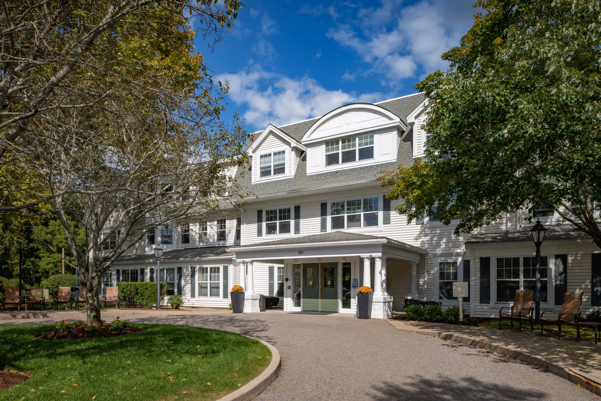 Front entrance of a white multi-story senior living building with columns, green double doors, and trees and chairs in the landscaped driveway.