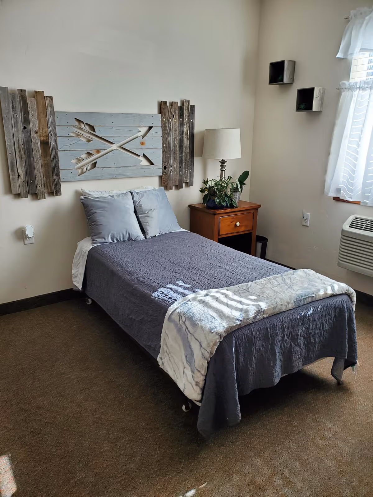 A neatly made single bed with gray bedding and two pillows in a small, cozy bedroom. Above the bed is a decorative wooden wall art featuring crossed arrows. To the right of the bed is a wooden nightstand with a lamp and a potted plant. A window with sheer white curtains allows natural light into the room, and an air conditioning unit is installed below the window.