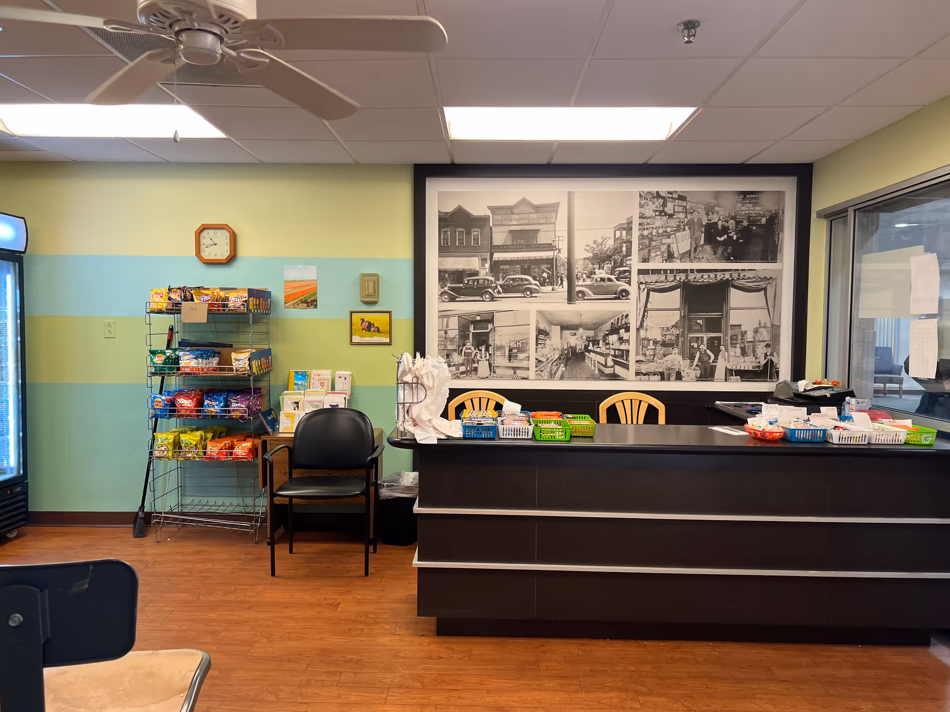 Interior view of a small snack shop area in Myers Apartments Independent Living, featuring a counter with baskets of snacks and items, a black chair, a rack filled with various bags of chips, a wall clock, and a large black and white historical photo collage on the wall behind the counter.