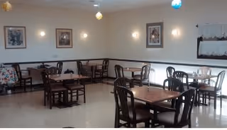 A dining room with several wooden tables and chairs arranged neatly. The walls are light-colored with framed artwork hanging, and there are small pendant lights hanging from the ceiling. A window with a white curtain is visible on the right side.