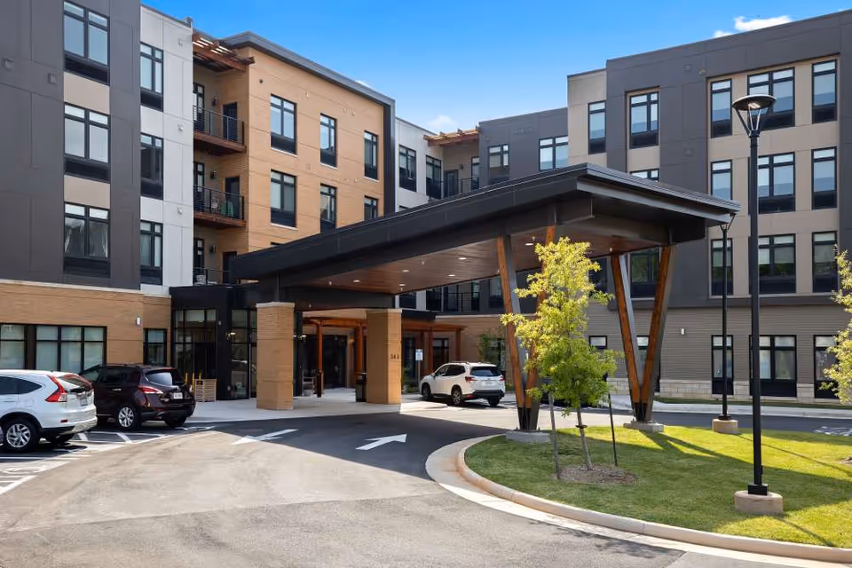 Exterior view of The Barclay at Charlottesville senior living facility showing a modern multi-story building with large windows, a covered entrance supported by wooden beams, parked cars, and a landscaped area with small trees and a street lamp under a clear blue sky.