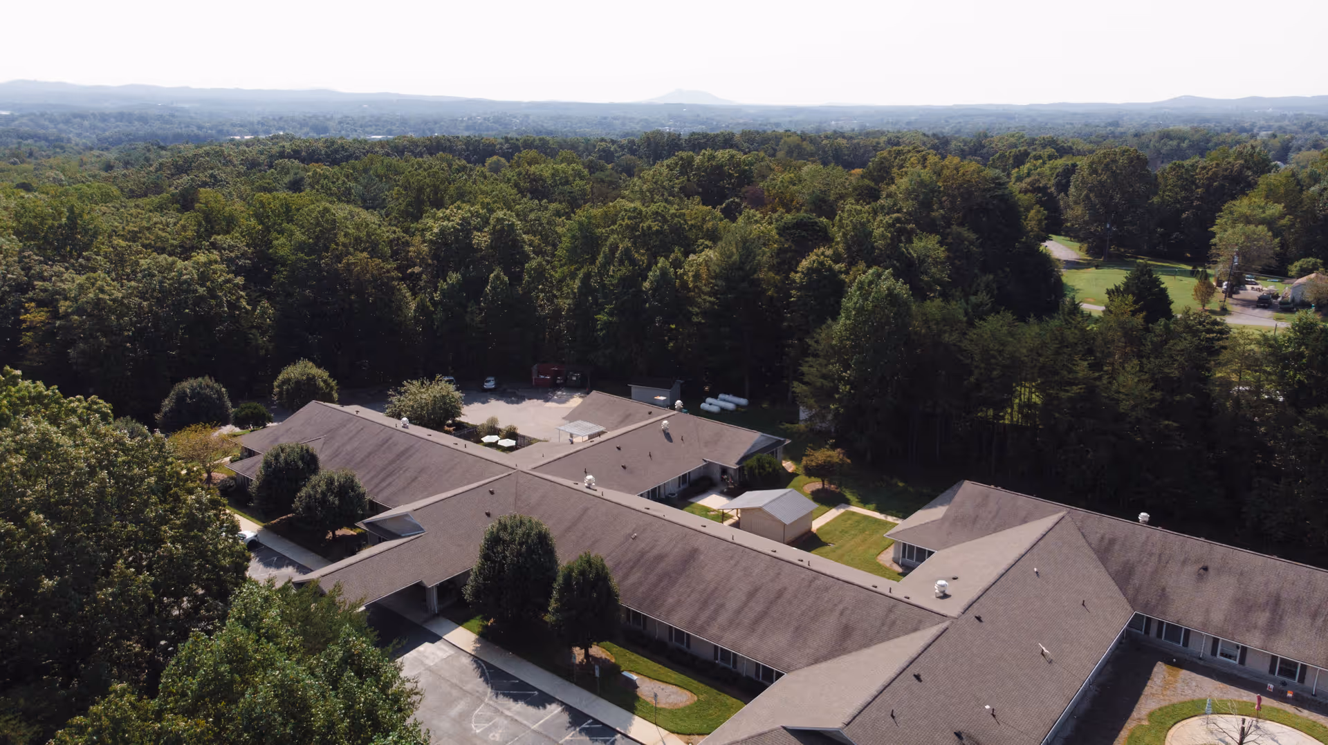 Aerial view of a senior living facility named Twelve Oaks surrounded by dense green trees and forested area, with a clear sky and distant hills in the background.