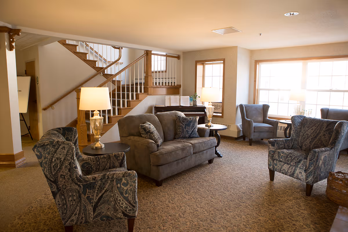 A cozy living room area with a gray loveseat, patterned armchairs, two side tables with lamps, large windows letting in natural light, and a wooden staircase in the background. There is also a piano against the wall with sheet music on it.