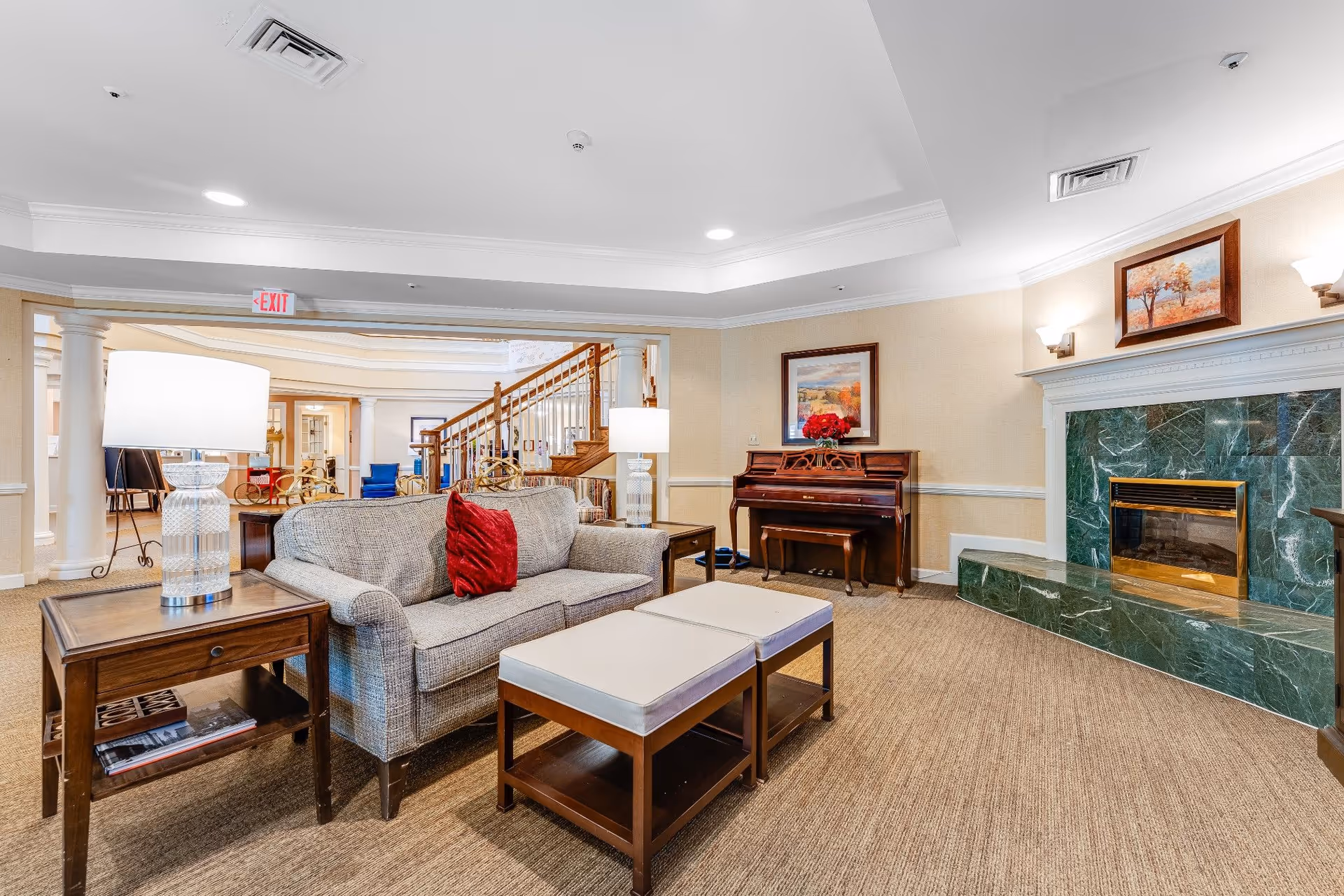 A cozy living room area in a senior living facility featuring a gray sofa with a red pillow, two cushioned ottomans, a wooden side table with a lamp, a green marble fireplace, and a wooden piano with a floral painting above it. The room has beige walls, carpeted floor, and recessed ceiling lights. In the background, there is a staircase and additional seating areas.