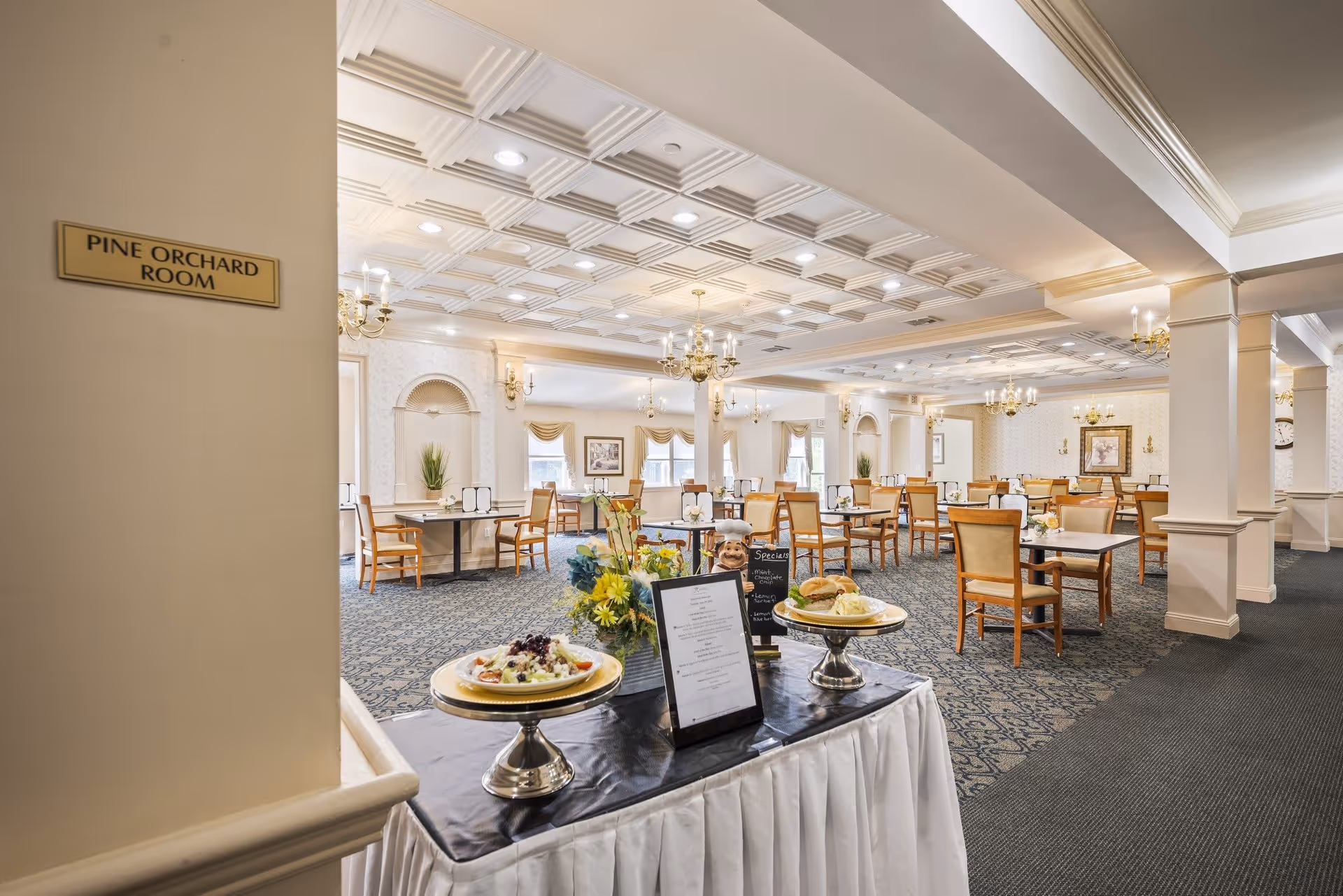 Interior view of the Pine Orchard Room dining area in HarborChase of Branford, featuring multiple tables and chairs arranged neatly on a patterned carpet. The room has a decorative ceiling with chandeliers, wall sconces, and large windows with curtains allowing natural light. In the foreground, a table displays plated food items, a menu, and a small chalkboard sign with specials.