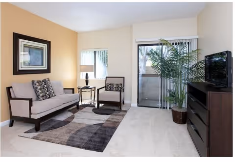 A bright living room with beige walls and carpet. The room features a light gray sofa with patterned cushions, a matching armchair, a round side table with a lamp, a large patterned area rug, a flat-screen TV on a dark wooden dresser, and a tall potted plant near sliding glass doors with vertical blinds leading to a balcony.
