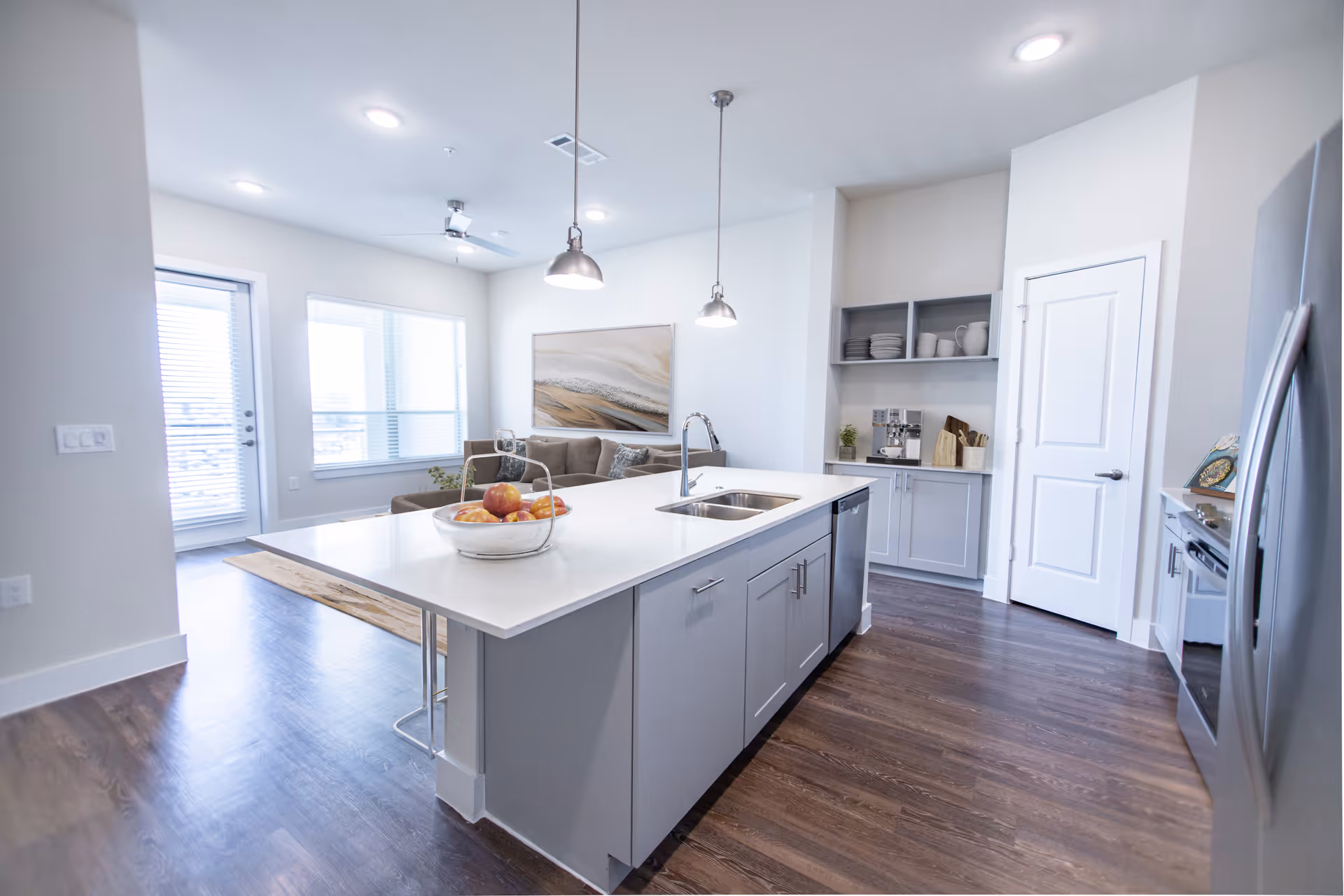 Bright modern open-plan kitchen with a large island, sink, pendant lights and a living area visible in the background.