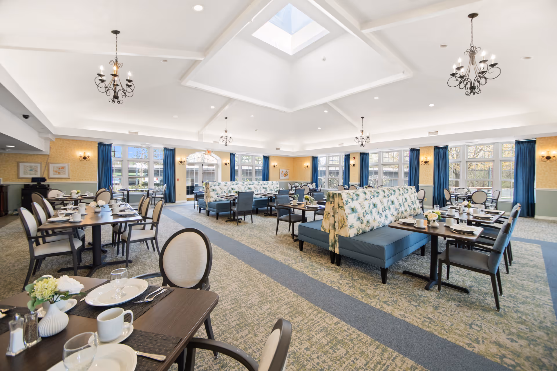 A spacious and well-lit dining room with multiple tables set with plates, cups, and silverware. The room features floral-patterned booth seating, upholstered chairs, large windows with blue curtains, chandeliers hanging from the ceiling, and a skylight allowing natural light to fill the space.