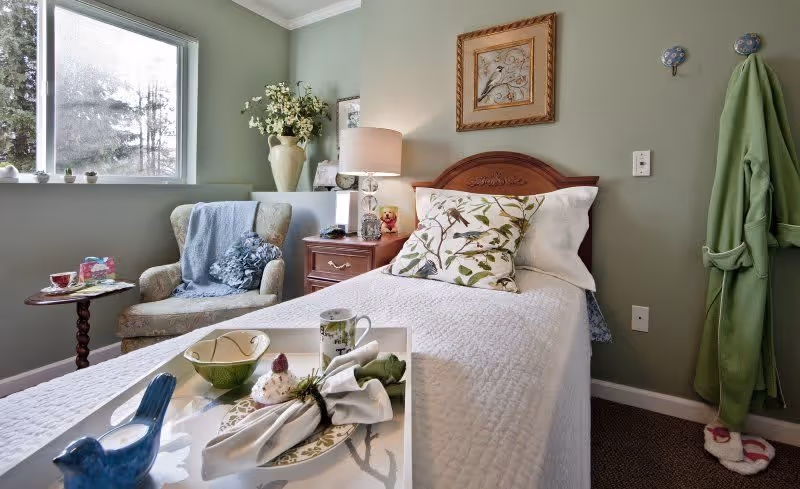A cozy bedroom with a single bed covered in a white quilt and a decorative pillow with a bird design. Next to the bed is a wooden nightstand with a lamp, a small teddy bear, and a vase with flowers. A comfortable armchair with a blue throw blanket and a small round table with a cup and a book are positioned near a window. A green robe hangs on the wall with slippers on the floor beneath it. The walls are painted light green and a framed bird artwork hangs above the bed.