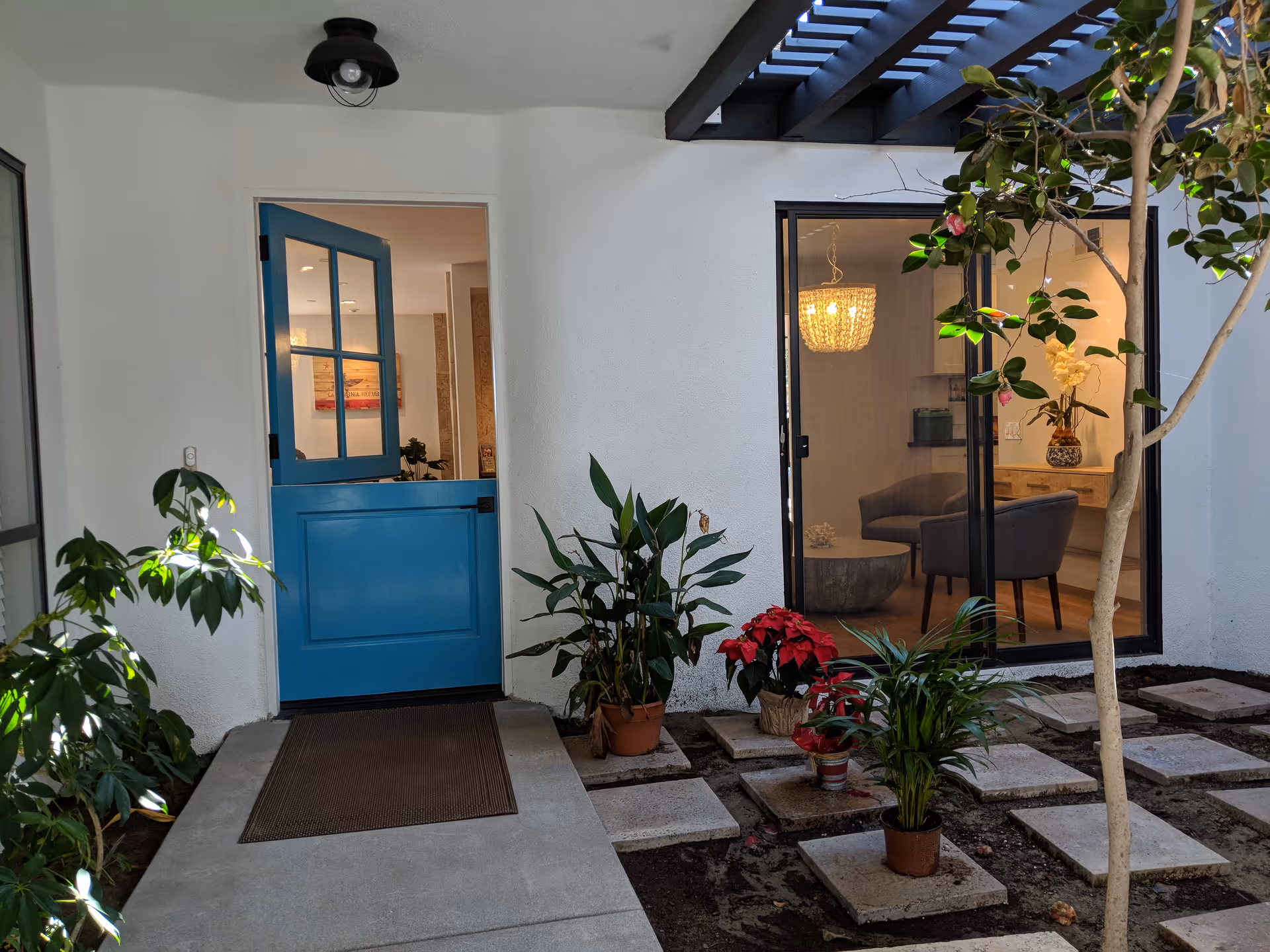 Small courtyard entrance with a blue Dutch door, potted plants on stepping stones, and a window showing a lit seating area inside.