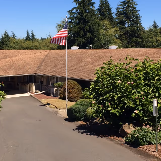 Exterior view of a single-story building with a brown roof surrounded by greenery and trees. An American flag is mounted on a flagpole near the driveway leading to the entrance of the building.