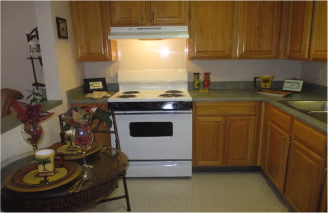 Small kitchen with a white electric stove, oak cabinets, green countertops, sink, and a small bistro table set for two.