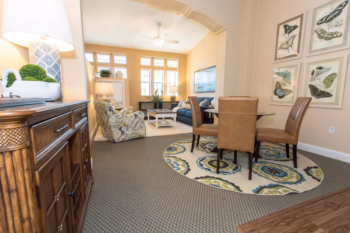 A cozy living room and dining area with beige walls and carpeted floor. The living room features a blue sofa, two patterned armchairs, a white coffee table, and two table lamps on side tables. The dining area has a round glass table with four brown leather chairs placed on a circular patterned rug. Butterfly artwork is displayed on the wall above the dining table.