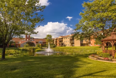 A scenic outdoor view of The Fountains at Atco featuring a grassy lawn, a pond with a water fountain, trees, a wooden gazebo on the right, and multi-story residential buildings in the background under a partly cloudy blue sky.