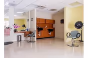 Interior view of a salon area in a senior living facility with two hair styling chairs, a hair dryer chair, a counter with a mirror, and a wooden cabinet mounted on a yellow wall.