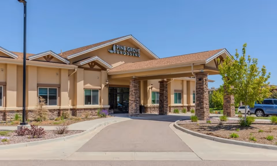 Exterior view of Pine Grove Crossing facility showing the entrance with a covered driveway supported by stone pillars, beige walls, and a clear blue sky.