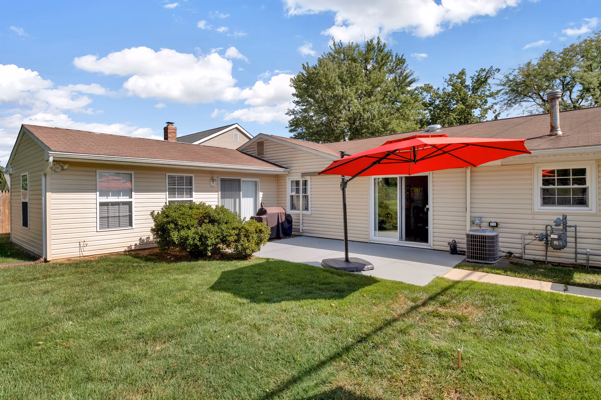 Backyard of a single-story beige house with a concrete patio, a large red umbrella providing shade, green grass lawn, bushes, and trees under a partly cloudy blue sky.