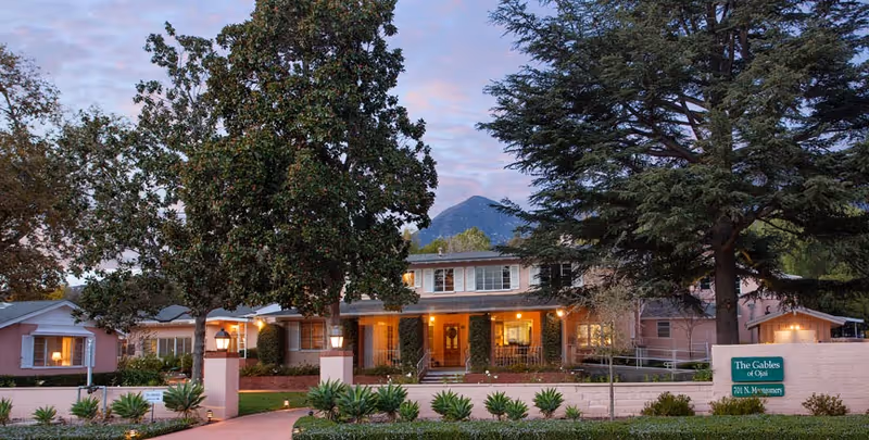 Exterior view of The Gables of Ojai senior living facility at dusk, showing a large two-story building with warm lights inside, surrounded by trees and landscaped greenery, with a mountain visible in the background.