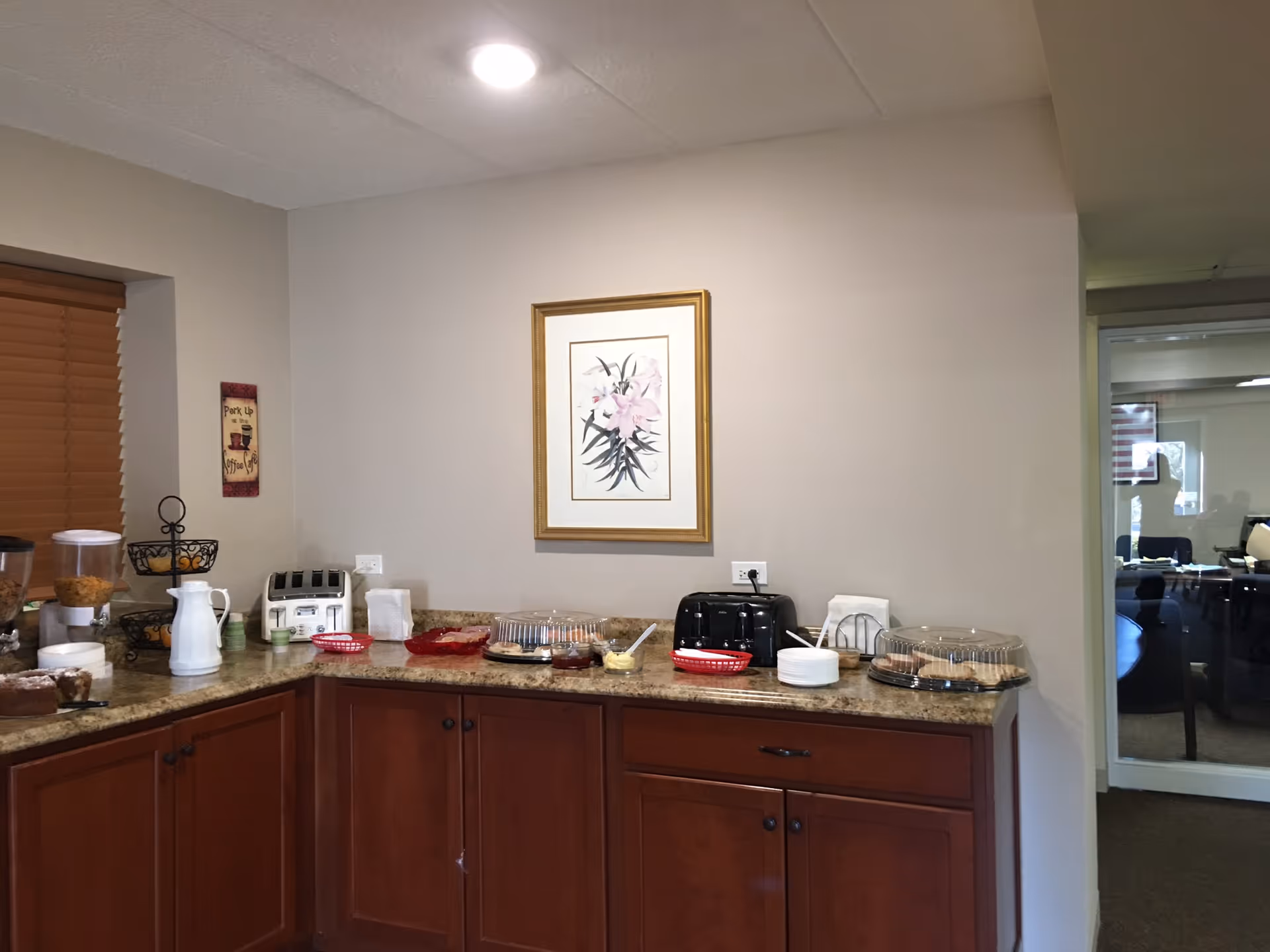 A small kitchenette/buffet area with a countertop, wooden cabinets, toaster, covered platters and framed floral artwork on the wall.