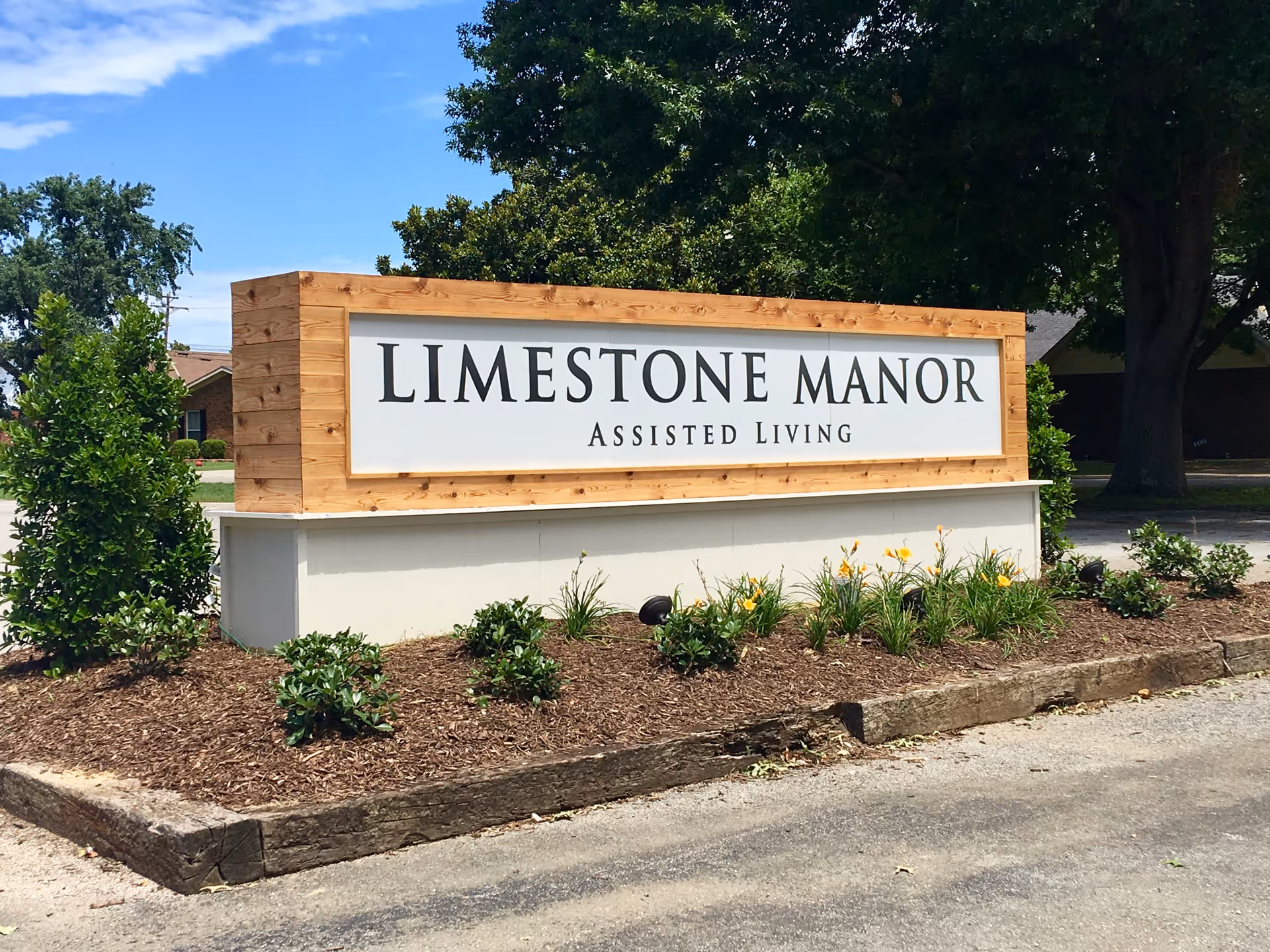 A wooden-framed roadside sign reading 'LIMESTONE MANOR ASSISTED LIVING' surrounded by mulch, plants, and trees.