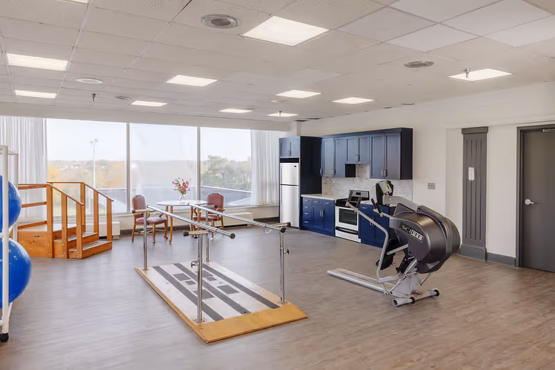 Spacious indoor rehab and activity room with parallel walking bars, exercise equipment, a kitchenette, seating, and large windows.