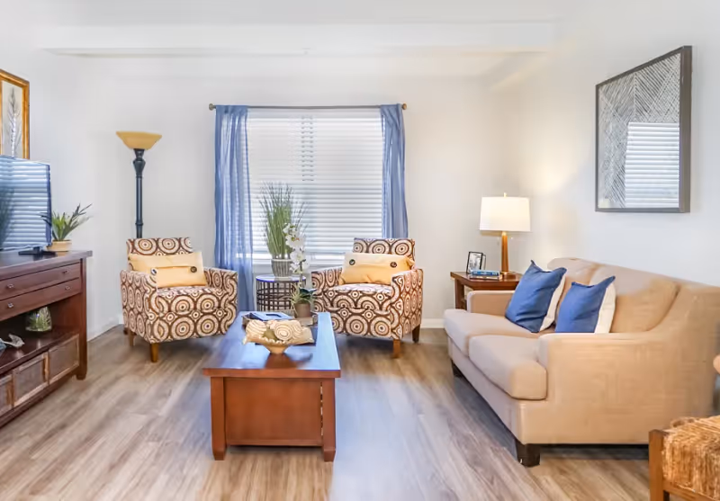 A bright living room with a beige sofa adorned with blue and white pillows, two patterned armchairs with yellow cushions, a wooden coffee table with decorative items, a wooden TV stand with a flat-screen TV, a floor lamp, and a window with blue curtains and white blinds.